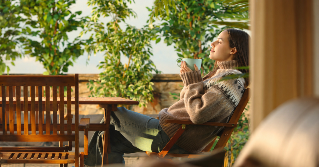 A woman enjoying afternoon sunlight and coffee while sitting on a teak outdoor chair on a terrace