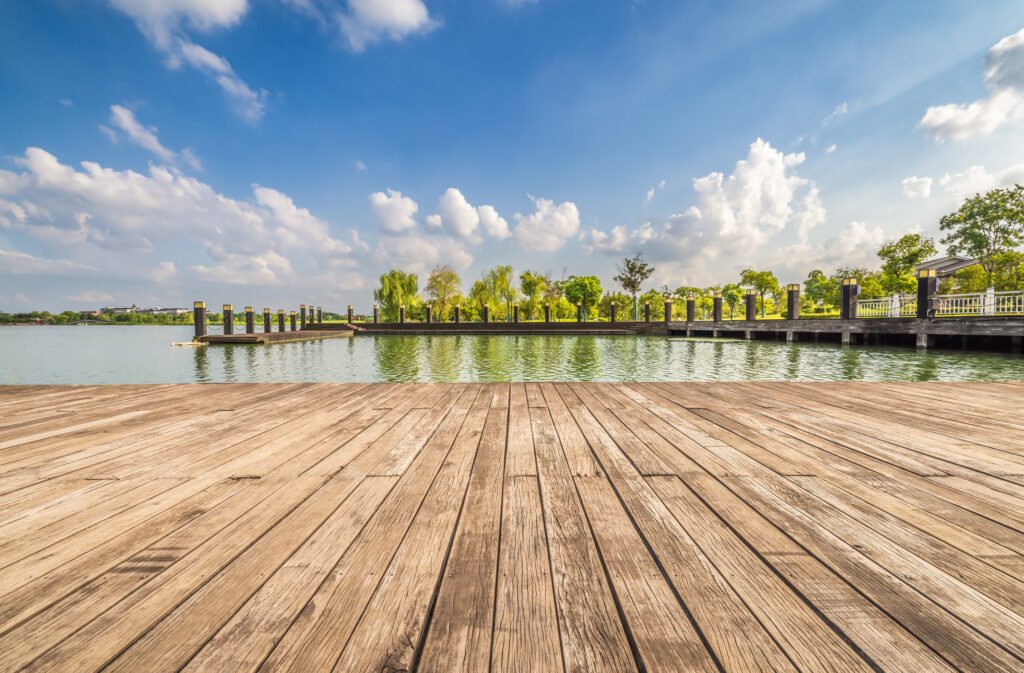 Relaxing wooden deck offers a tranquil view of the lake and lush trees, all under the vast expanse of a clear blue sky. Waterfront outdoor deck exposed to sun and moisture, highlighting environmental factors that affect outdoor furniture durability.