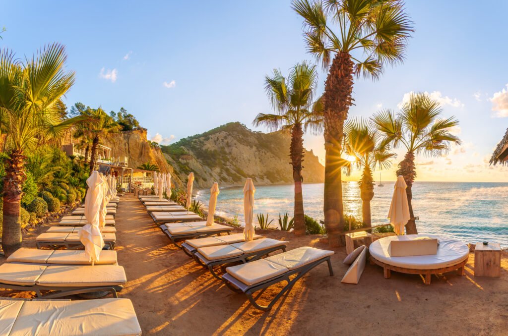 Luxury beachfront resort with outdoor sun loungers, umbrellas, palm trees, and a round daybed overlooking the ocean at sunset.