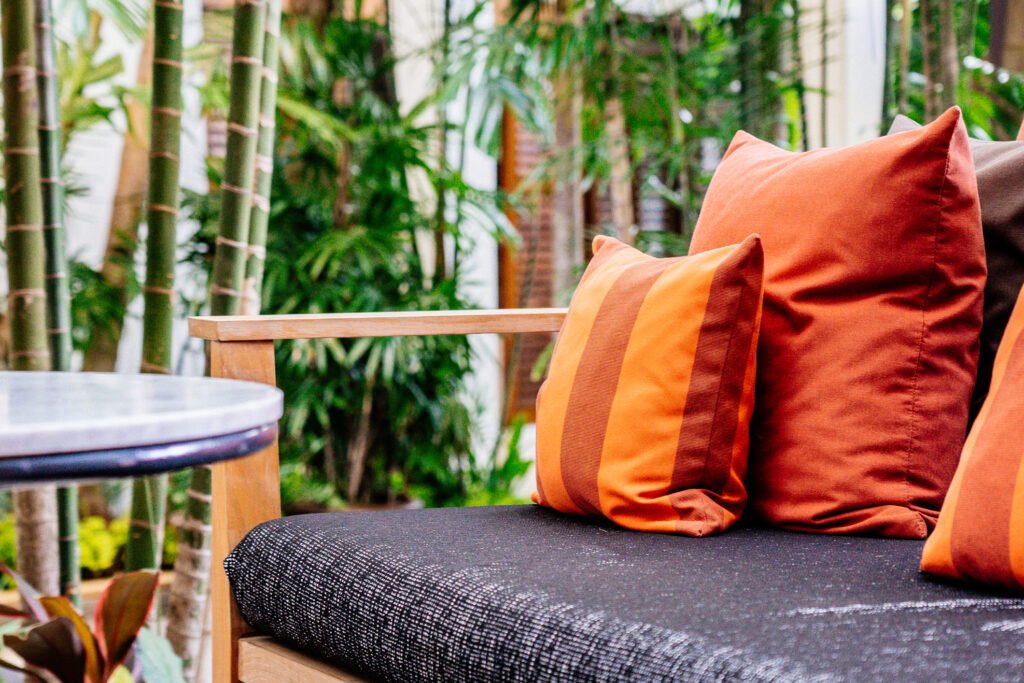 Close-up of a wooden outdoor lounge chair with dark seat cushion and orange throw pillows in a lush tropical garden setting.
